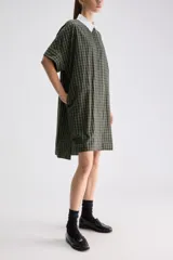 Vestido camisero midi de calce relajado a cuadros en tonos verde y negro, confeccionado en algodón liviano. Presenta cuello clásico en contraste de color blanco, cartera frontal oculta con botones y mangas cortas con vuelta.