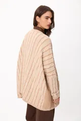 Sweater de punto con diseño de trenzas verticales, color crudo y camel, con bolsillos frontales y detalle de logo bordado. Presenta un corte holgado y mangas largas.