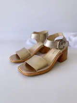 Sandalias de cuero color dorado, con tira ancha sobre los dedos, pulsera al tobillo con hebilla metálica y taco de madera.