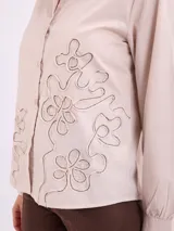 Camisa beige de manga larga con cuello camisero, cierre frontal con botones y diseño de pespuntes y bordados en relieve con formas florales en la parte inferior y lateral.