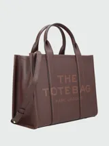 Cartera tipo tote de cuero granulado color tostado, con diseño estructurado y rectangular. Presenta doble asa superior fija y una correa larga ajustable y extraíble. En el frente, destaca el logo estampado con la inscripción 'THE TOTE BAG MARC JACOBS'.