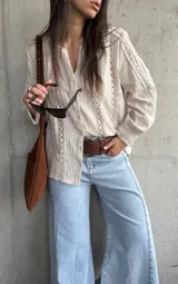 Camisa de manga larga color beige con detalles de encaje y calados verticales en el frente y las mangas, cuello en V y cierre frontal con botones.