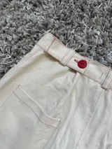 Bermuda de jean color crudo con costuras y detalles en rojo, incluyendo un botón frontal y un logo bordado en el bolsillo lateral. Presenta un corte recto y largo hasta la rodilla.