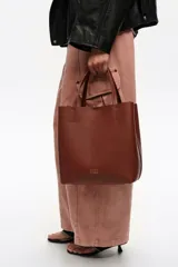 Bolso de hombro de tamaño mediano color marrón, confeccionado en piel vacuna. Presenta doble asa corta de mano y correa de tejido jaspeada con el nombre de la marca estampado. Incluye detalles en piel, cierre con botón de presión, logo grabado en dorado en el frente y acabados metálicos.