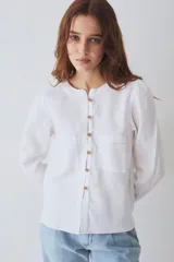 Blusa de lino color blanco, con cuello redondo, manga larga y cierre frontal con botones de madera. Presenta dos bolsillos frontales tipo parche con costuras a la vista.