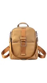 Mochila de tamaño mediano, color beige claro, con cuerpo principal de tela impermeable y detalles en simil cuero liso en la parte inferior y correas. Presenta un asa superior corta y correas ajustables para llevar al hombro o cruzada. Cuenta con un bolsillo frontal con cierre y un detalle decorativo de correa con hebilla metálica rectangular.