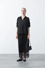 Camisa negra de corte relajado y caída fluida, con cuello clásico, escote en V y cierre frontal con botones forrados. Presenta mangas largas con puños abotonados.
