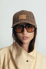 Gorra tipo baseball con estampado de cuadros escoceses en tonos azul, naranja y beige. Presenta un parche de cuero sintético en el frente y cierre ajustable en la parte posterior.