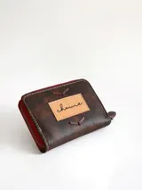 Billetera pequeña de cuero color chocolate con cierre perimetral rojo y detalle de costuras decorativas en forma de espiga en el frente.