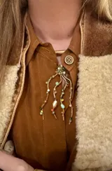 Collar de bronce con múltiples tiras colgantes decoradas con cuentas de colores y piedras, complementado con tiento marrón.