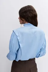 Blusa celeste de manga larga con puños fruncidos y elásticos, escote en V con cuello alto con volados y delicados bordados florales en el pecho.