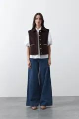 Chaleco de pana color marrón oscuro, con cuello camisero, cierre frontal de botones metálicos dorados y dos bolsillos frontales con detalle de bordado geométrico.