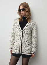 Cardigan de punto grueso con diseño jaspeado en blanco y negro. Presenta cuello en pico, cierre frontal con botones efecto joya, dos bolsillos de parche delanteros y detalles en contraste negro en los bordes.