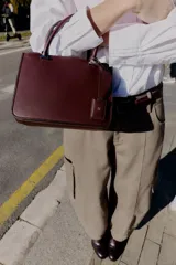 Bolso pequeño tipo satchel, color borgoña oscuro, con doble asa de mano y correa bandolera extraíble. Presenta dos compartimentos exteriores con cierre de cremallera metálica y un compartimento interior con cierre imantado.