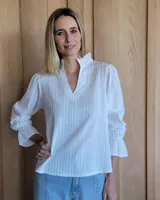 Blusa blanca de algodón con textura de rayas verticales caladas, cuello con volados y escote en V. Presenta mangas largas con puños acampanados y volados.