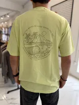 Remera oversize color verde claro con un gran estampado gráfico en la espalda de estilo tropical que incluye palmeras, olas, sol y texto circular.