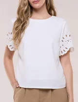 Blusa blanca de corte recto con cuello redondo y mangas cortas que presentan detalles de bordado calado y terminación festoneada.