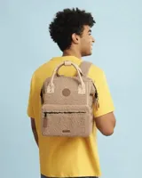 Mochila Cabaia modelo Adventurer Manchester, confeccionada en tejido tipo corderito color beige. Presenta asas superiores y detalles en cuero sintético color crema, bolsillo frontal con cierre y parche de la marca en el centro.