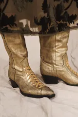 Botín de piel estilo cowboy con acabado metalizado dorado, cordones en el empeine, tacón medio ancho asimétrico y puntera perfilada.
