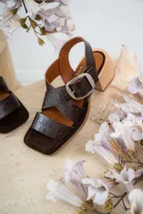 Sandalias de cuero negro con textura de reptil, diseño de punta cuadrada y taco bloque de madera. Presentan tiras cruzadas en el empeine y una hebilla metálica grande decorativa.
