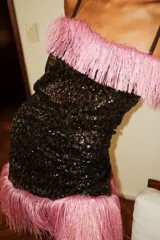 Vestido corto sin mangas, color negro, completamente cubierto de lentejuelas brillantes. Presenta un borde de flecos largos de color rosado en el escote y en el ruedo inferior.