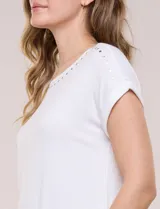 Remera blanca de punto acanalado con escote redondo adornado con tachas metálicas en el cuello y hombros, mangas cortas japonesas y ruedo curvo.
