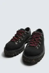 Zapato de cordones estilo trekking para hombre, confeccionado en piel con acabado de serraje color marrón. Presenta enganches metálicos para los cordones, cuello acolchado en color negro y una suela de goma con volumen y efecto moteado.