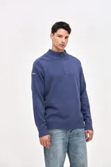 Buzo de punto color azul, con cuello alto y cierre frontal parcial. Presenta un diseño con textura acanalada en los hombros y mangas, y un pequeño bordado en la manga derecha.