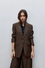 Blazer de silueta amplia y largo por debajo de la cadera, confeccionado en tela liviana con brillo sutil y caída fluida color marrón. Presenta cuello con solapas, cierre frontal, bolsillos con tapa, mangas largas y abertura posterior.