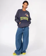 Buzo de algodón y poliéster, color azul oscuro con efecto desgastado/tie-dye. Presenta un gran estampado frontal con la palabra "MICHIGAN" en amarillo con borde negro, un corazón naranja y apliques de pines decorativos en la parte superior. Tiene cuello redondo con terminación deshilachada y bolsillo canguro frontal.