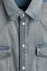 Camisa de denim de algodón rígido, de corte oversized y silueta holgada. Cuenta con cuello clásico, cierre frontal de botones a presión, canesú en la parte delantera y trasera, bolsillos con solapa en el pecho, mangas largas con puños abotonados y bajo redondeado.