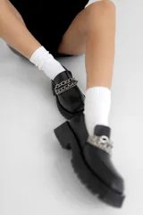 Mocasín de cuero color suela con punta cuadrada y suela track de goma oscura. Presenta un detalle de doble cadena metálica plateada sobre el empeine.