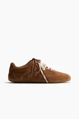 Calzado deportivo de gamuza suave en color marrón, con diseño de perfil bajo, collarín acolchado y doble juego de cordones en tonos marrón y blanco.