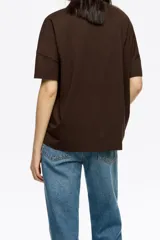 Camiseta de manga corta, corte oversize, color marrón oscuro. Presenta un estampado frontal con el texto "BIMBA Y LOLA" realizado con hilos de colores vibrantes que cuelgan, simulando un bordado texturizado.