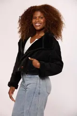 Campera corta tipo gamulán en color negro, con interior y detalles en puños y cuello de corderito sintético al tono. Presenta cierre frontal metálico y un botón decorativo en el ruedo.