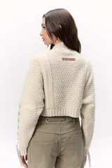 Sweater de lana merino tejido en punto grueso, color gris oscuro, con cuello alto. Presenta un diseño calado con tientos de cuero marrón insertados a lo largo de la prenda, simulando un lazo o detalle orgánico.