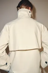 Trench corto color marfil de algodón, con cuello camisero, solapa cortaventos, mangas largas amplias con puños abotonados y cierre cruzado con un botón visible.