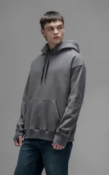 Hoodie oversize color gris oscuro, confeccionado en frisa premium con textura gamuzada. Presenta capucha ajustable con cordones personalizados, bolsillo frontal tipo canguro y puños con abertura para el pulgar.