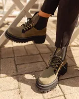 Borcego de gamuza color chocolate con paneles laterales elásticos negros y suela track dentada. Presenta sistema de cordones con ganchos metálicos y tirador en la parte trasera.
