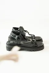 Sandalias negras de estilo chunky con plataforma dentada. Presentan tiras ajustables con cierre de velcro y hebillas metálicas, combinando texturas de cuero granulado y detalles en charol.