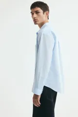 Camisa en mezcla de lino y algodón color celeste, de corte relajado, manga larga, con cuello inglés, bolsillo frontal y cierre de botones.