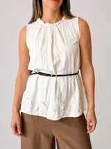 Blusa blanca sin mangas, con cuello trenzado fruncido, detalles de volados en el ruedo y un cinturón fino marrón incorporado.