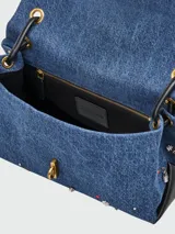 Bolso de hombro rectangular de denim azul, con solapa frontal decorada con múltiples charms metálicos y de esmalte, incluyendo cerezas, lazos, estrellas y candados. El bolso tiene herrajes dorados y correa de hombro de cuero negro.