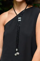 Collar largo tipo lazo con cuentas negras cilíndricas, detalle de pieza metálica central con incrustaciones y terminaciones con dijes metálicos circulares texturizados.