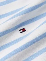 Chomba de piqué de algodón elástico, color blanco con rayas horizontales celestes. Presenta cuello polo blanco con ribete rojo y azul, tapeta de botones y logo de Tommy Hilfiger bordado en el pecho.
