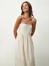 Vestido midi color beige claro, de corte amplio y fluido, con tirantes finos ajustables mediante lazos en los hombros, escote recto y prácticos bolsillos laterales.