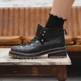 Borcego de cuero negro de estilo montañés con detalles en marrón oscuro y suela dentada de goma en tono caramelo. Cuenta con cordones negros y ganchos metálicos.
