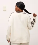 Cardigan deportivo de corte oversized, color blanco con detalles en marrón oscuro. Presenta escote en V, cierre frontal con botones a presión, mangas largas con franjas laterales y puños acanalados. Incluye un parche circular con letras en el pecho.