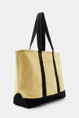 Bolso shopper de lona color beige con base, asas y tiras frontales en color negro. Cuenta con un bolsillo frontal con botón a presión y costuras visibles en contraste.