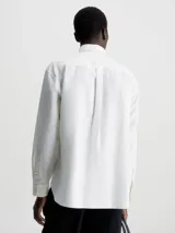 Camisa blanca de manga larga con cuello clásico, cierre frontal con botones, bolsillo de parche en el pecho y logo CK bordado en el bolsillo.